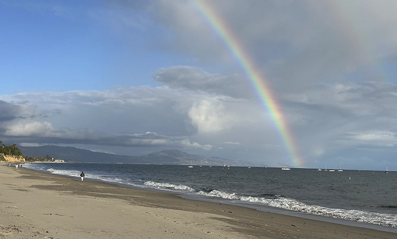 Rainbow arching over a quiet Santa Barbara beach with dramatic clouds and blue sky