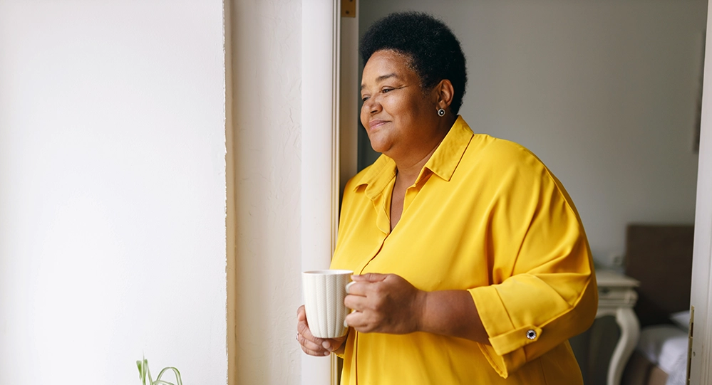 Woman holding a cup of tea and looking out a window with a peaceful expression 