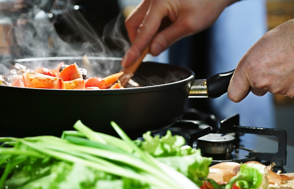 Hands stirring vegetables in a steaming skillet with fresh greens nearby
