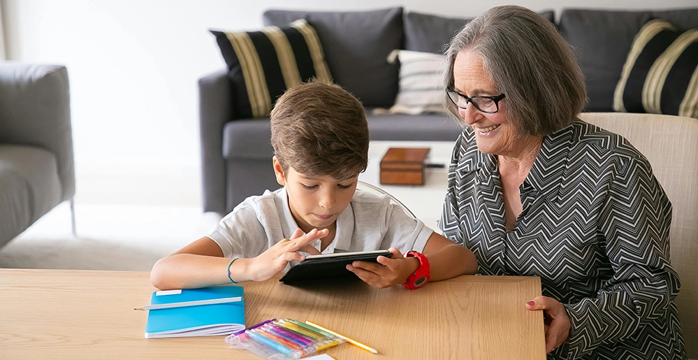 Grandmother and grandson working together on a tablet with homework supplies nearby — the kind of everyday caregiving that boosts brain health