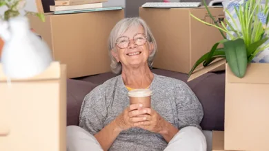 Senior woman with silver hair and glasses sitting contentedly among moving boxes in her new home, holding a coffee cup — moving after 50 with ease and grace