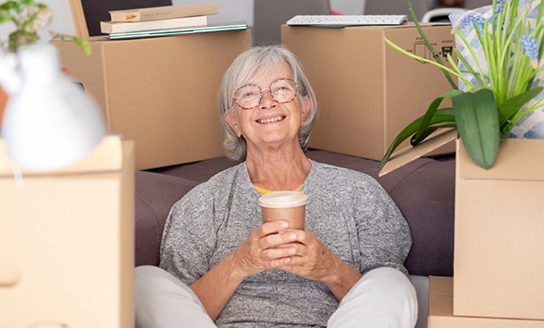 Senior woman with silver hair and glasses sitting contentedly among moving boxes in her new home, holding a coffee cup — moving after 50 with ease and grace