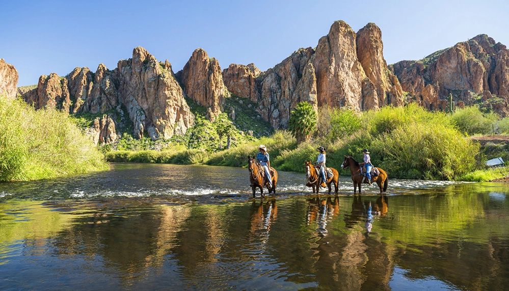 Horseback riders crossing a desert river beneath dramatic red rock canyon walls in Scottsdale's Sonoran Desert