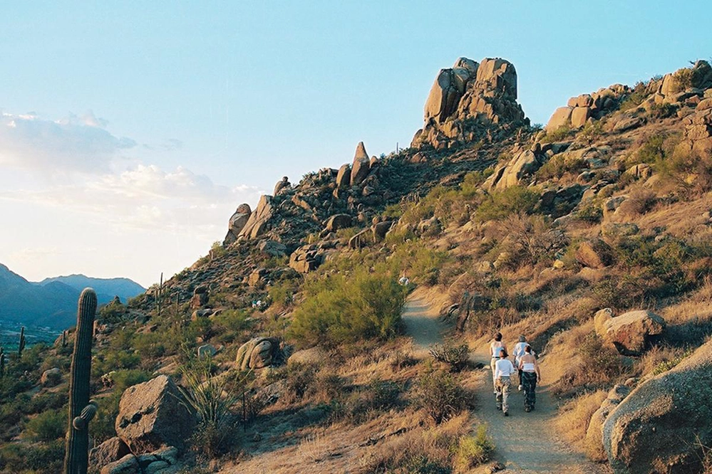 Hikers walking a desert trail toward Pinnacle Peak at sunset with saguaros and rock formations