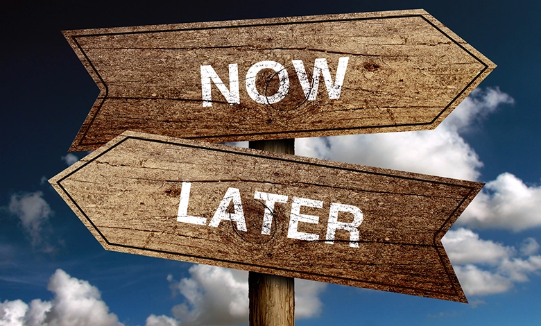 Wooden directional signs reading Now and Later pointing opposite directions against a blue sky