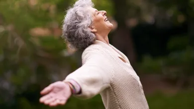 Older woman with arms outstretched and head tilted back in joyful laughter, outdoors among greenery