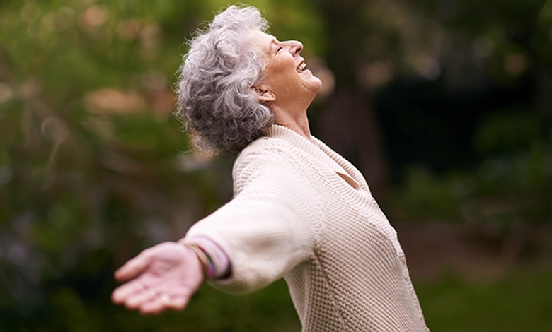 Older woman with arms outstretched and head tilted back in joyful laughter, outdoors among greenery