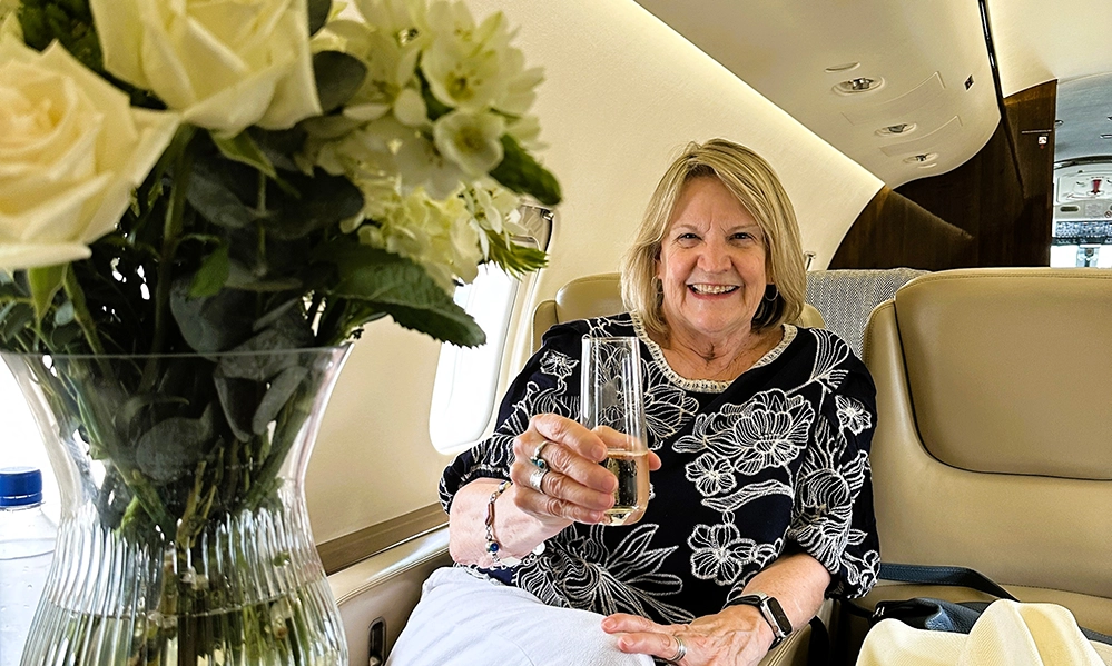 Barbara Redding raising a glass of champagne in a private jet cabin, with white roses on the table beside her.