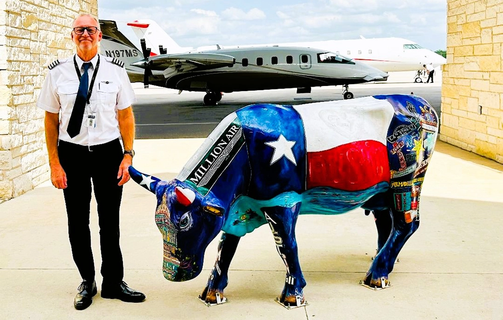 Cirrus Aviation pilot Philip Crossley standing beside a Texas-flag-painted cow statue with a private jet at the Million Air Austin terminal.