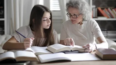Grandmother with glasses helping teenage granddaughter with homework at a desk surrounded by bookshelves
