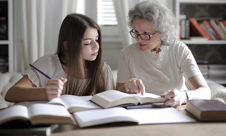 Grandmother with glasses helping teenage granddaughter with homework at a desk surrounded by bookshelves