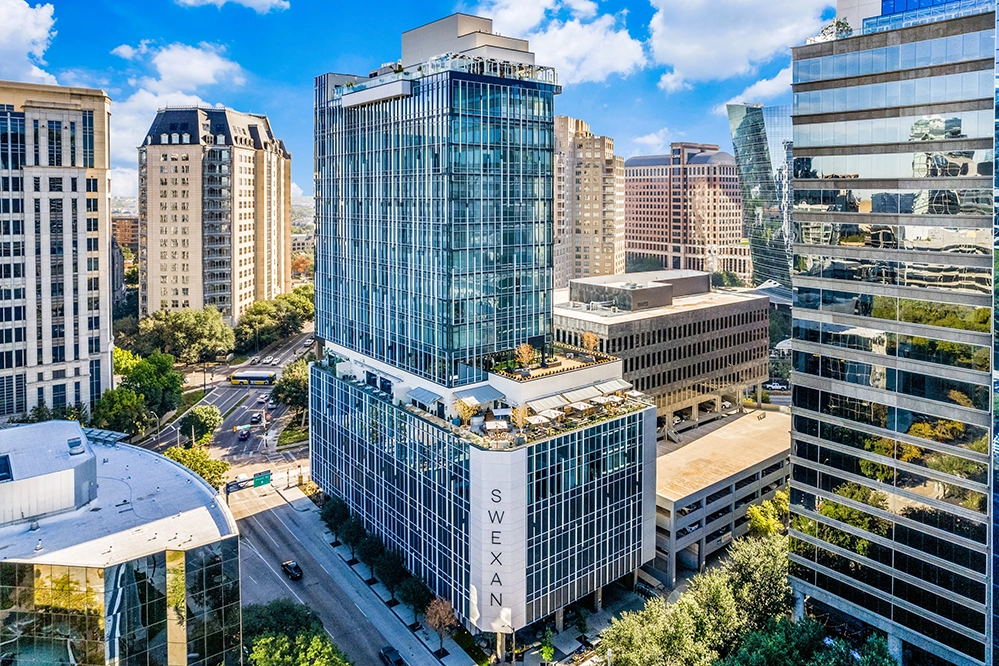 Exterior view of the 22-story Hôtel Swexan in Dallas, with SWEXAN signage and rooftop terraces visible above the Harwood District.