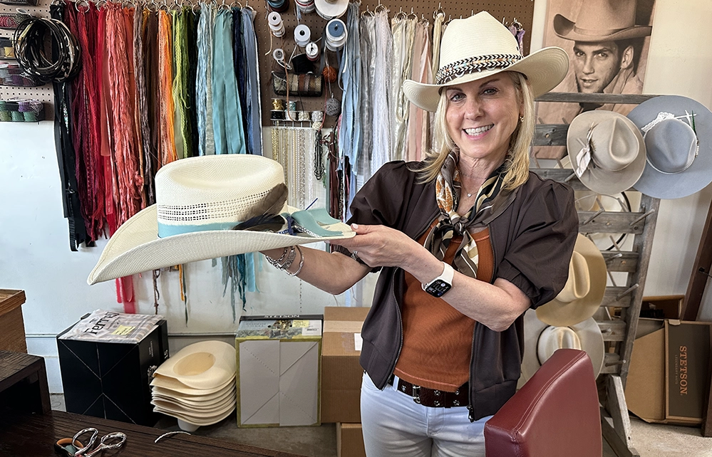 A McKinney Hat Company stylist wearing a Stetson cowboy hat holds another customized straw Stetson with ribbons and feathers at the hat company workshop.