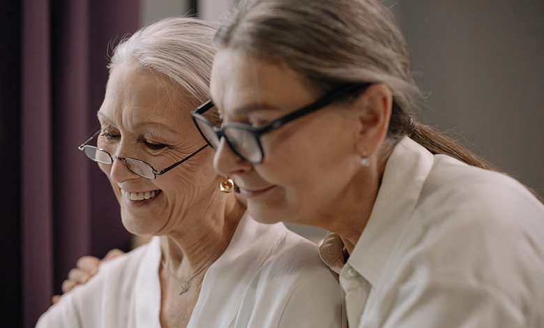 Two older women smile warmly together, one resting a hand on the other’s shoulder, illustrating the connection that endures through the dementia journey.