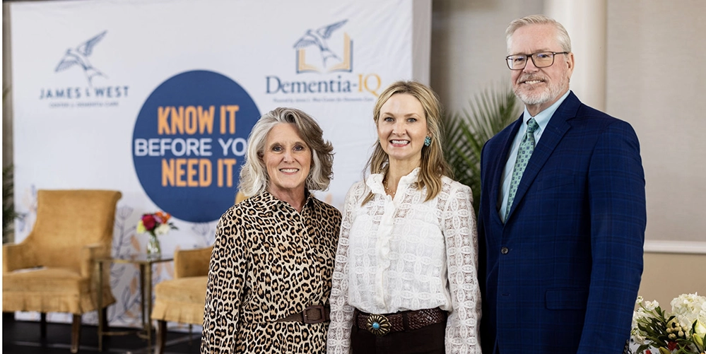 Three guests stand together in front of the “Know It Before You Need It” event backdrop.