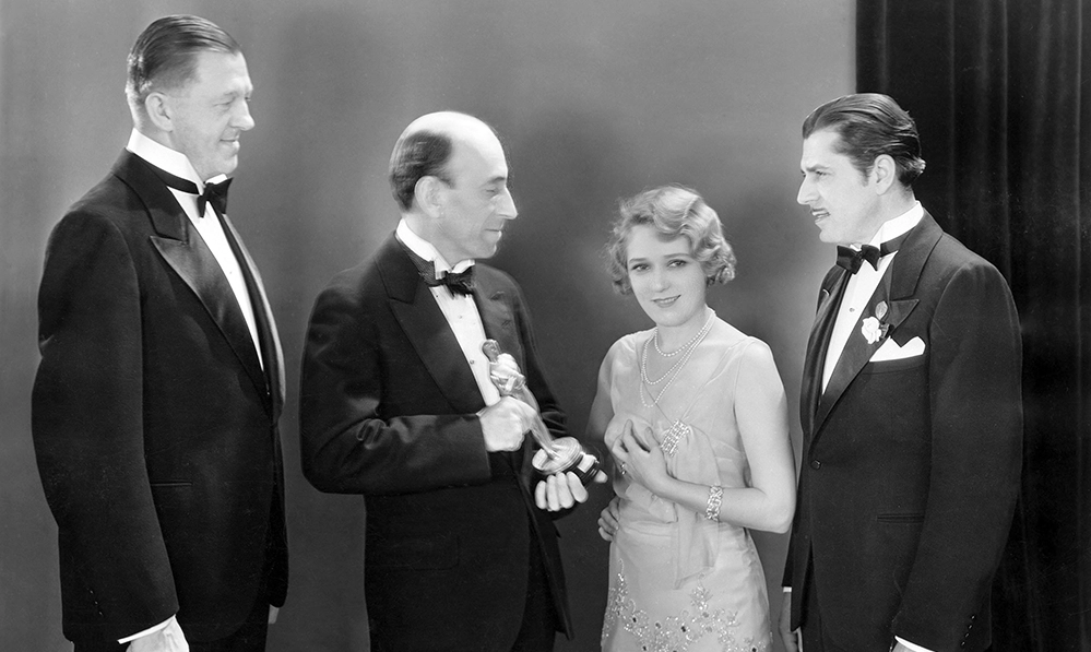 Black and white photograph of Mary Pickford receiving her Academy Award for Best Actress for Coquette at the 1930 ceremony surrounded by three men in formal attire