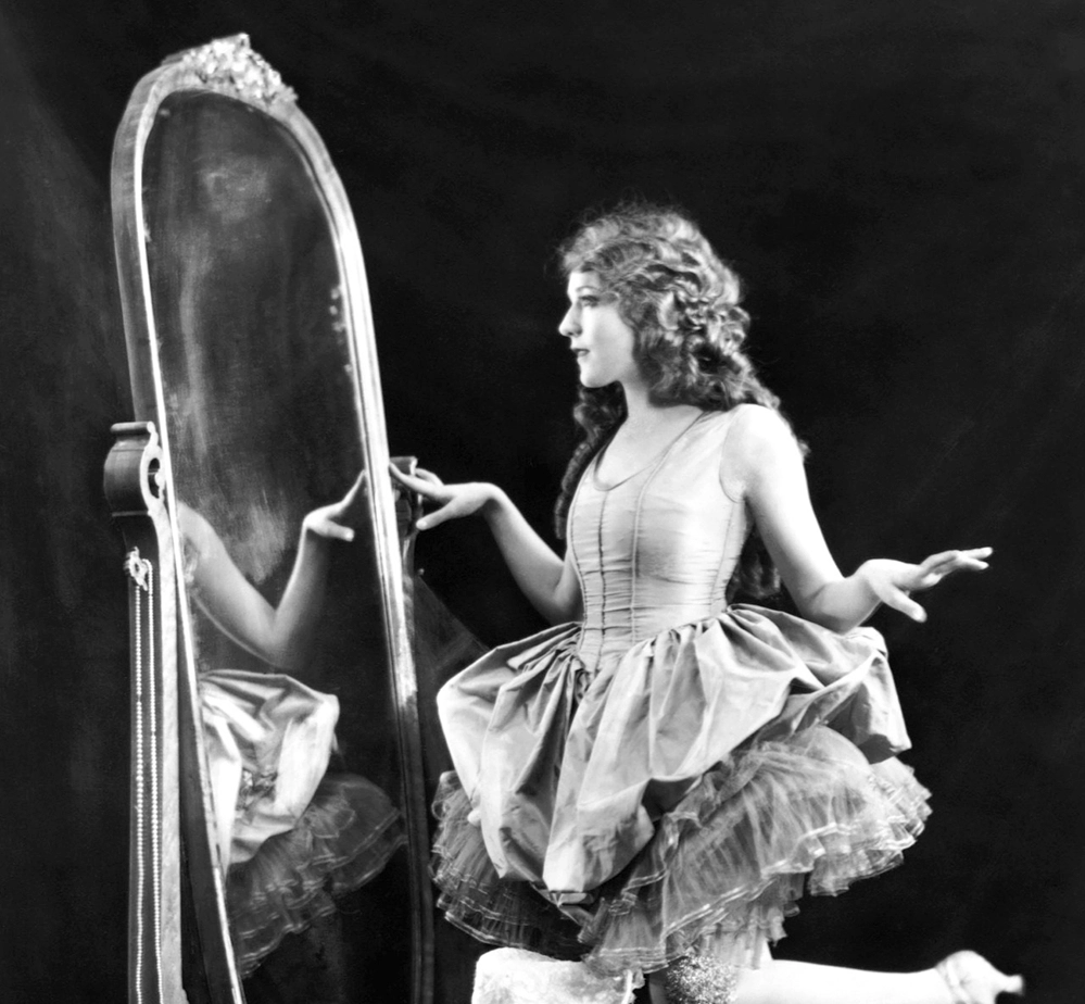 Black and white photograph of Mary Pickford in a ruffled dress seated on a stool beside a full length mirror her reflection visible