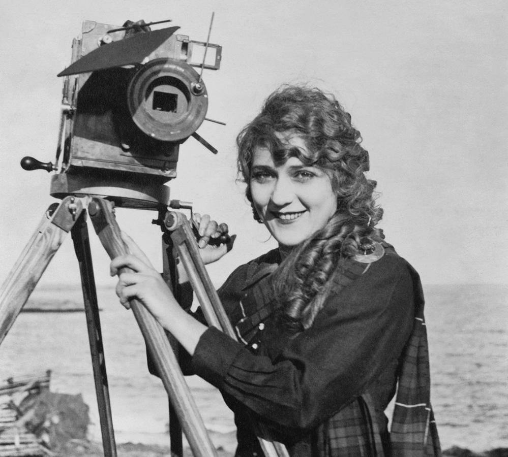 Black and white photograph of Mary Pickford smiling while standing beside a large film camera on a tripod outdoors by the ocean