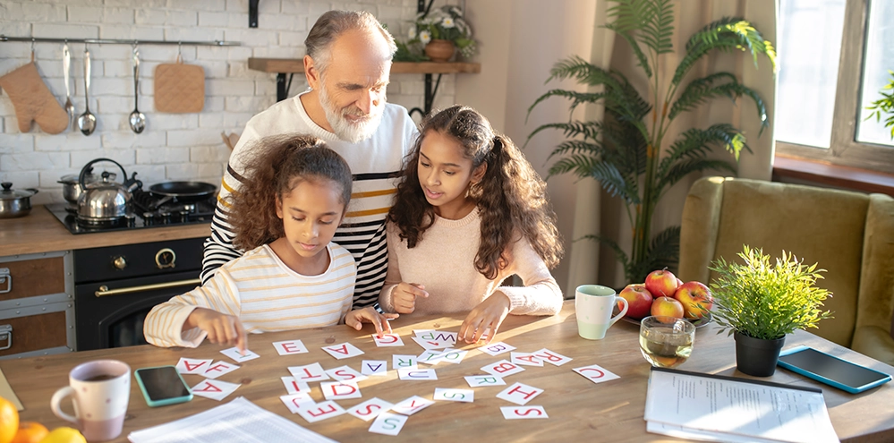 Memory Exercises for Sharper Recall After 50: 3 Levels to Train Your Brain 5 Grandfather and granddaughters arranging letter cards at the kitchen counter — a family memory exercise for all ages.