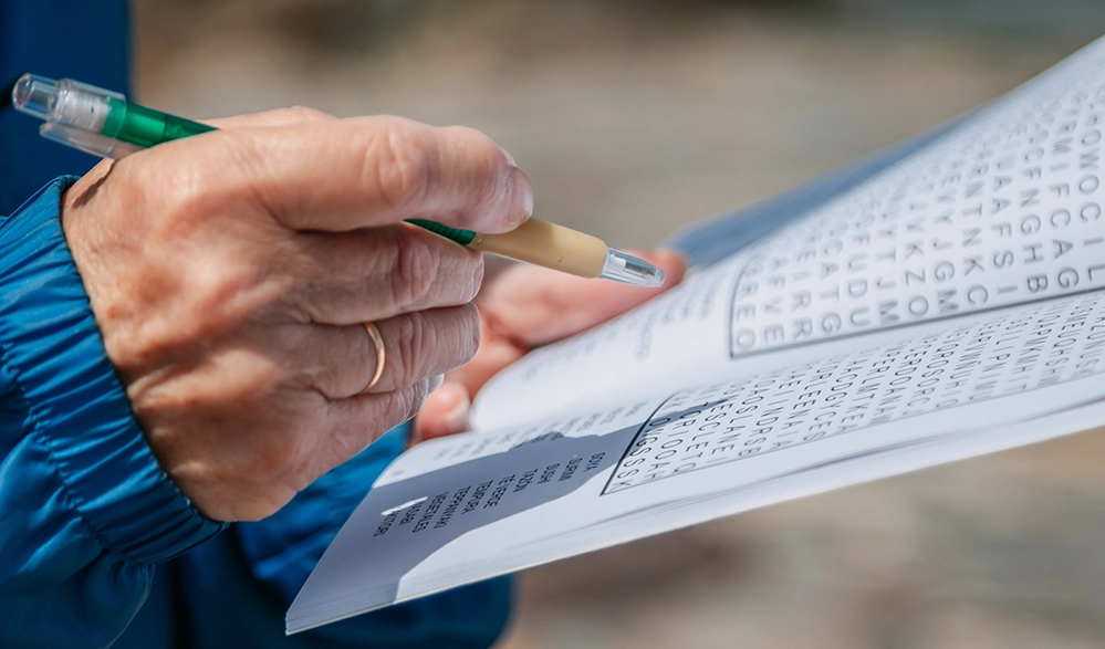 Memory Exercises for Sharper Recall After 50: 3 Levels to Train Your Brain 3 Senior hands holding a word search booklet with a pencil mid-stroke — a quiet memory exercise after 50.