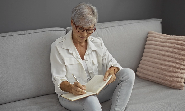 An older woman sitting on a couch, writing in a notebook with focused concentration — a daily memory exercise after 50.