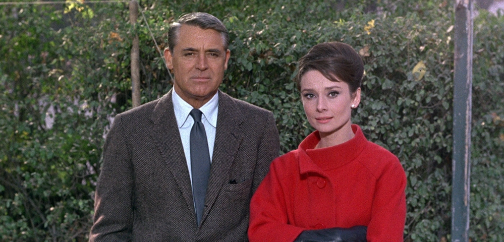 Cary Grant in a tweed jacket beside Audrey Hepburn in a red coat in the 1963 film Charade.