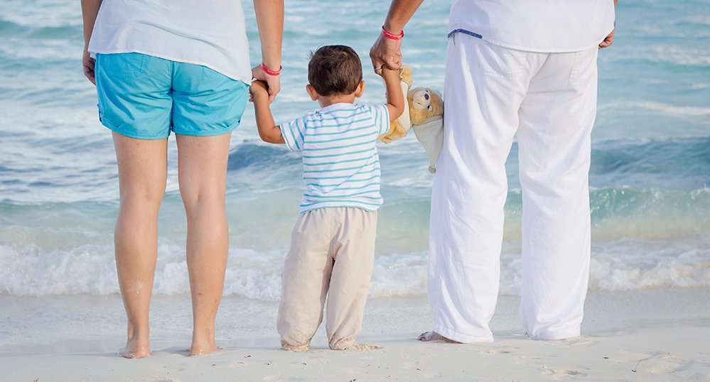 A young child holding hands with two adults at the ocean’s edge