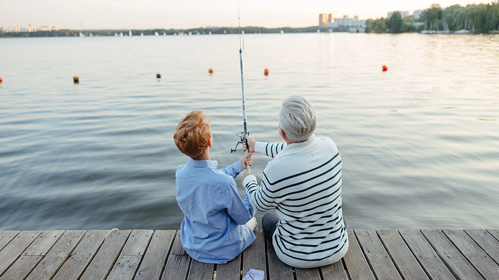 A grandfather and his grandson fishing together on a lake dock at sunset 