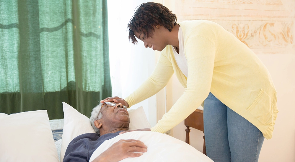 Caring for Aging Parents at Home: A Practical Guide for Adult Children 3 A woman gently tends to an older man resting in bed at home, reflecting the hands-on reality of home care for aging parents.