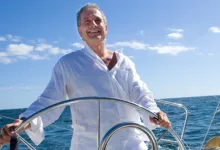 A smiling older man steering a sailboat on open water under a bright blue sky.