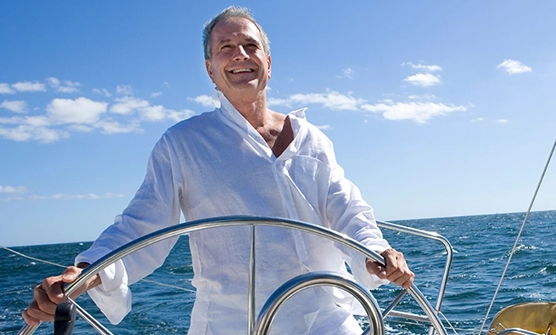 A smiling older man steering a sailboat on open water under a bright blue sky.