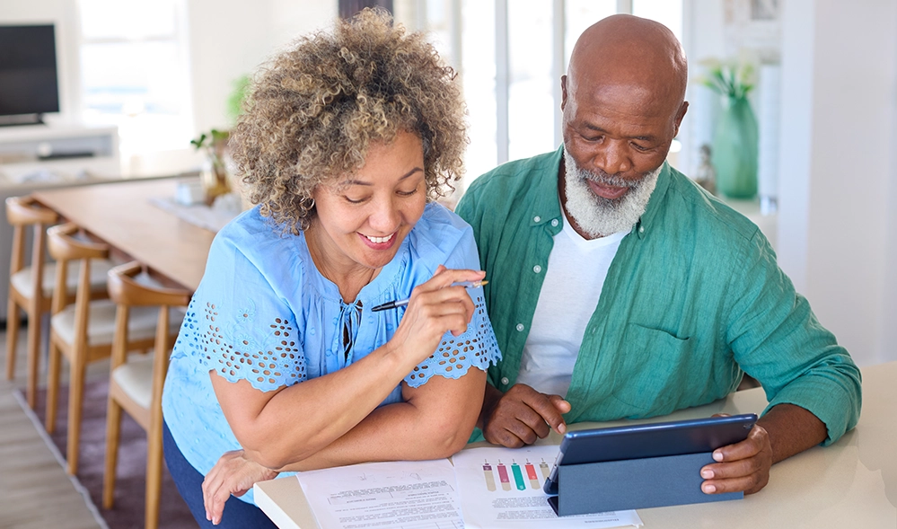 An older couple at a kitchen counter reviewing financial documents and a tablet together