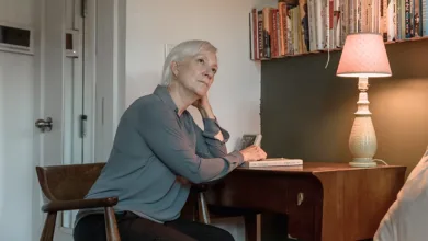 An older woman sits at a desk beside a lit lamp and a wall of books, resting her chin on her hand, an unopened book in front of her.