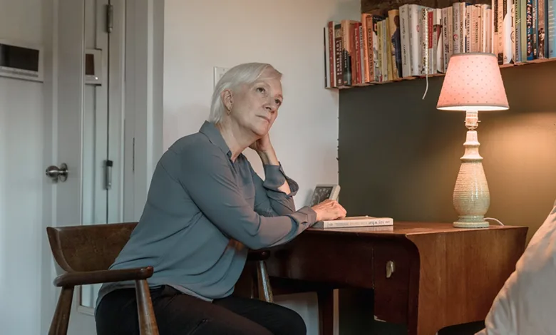 An older woman sits at a desk beside a lit lamp and a wall of books, resting her chin on her hand, an unopened book in front of her.