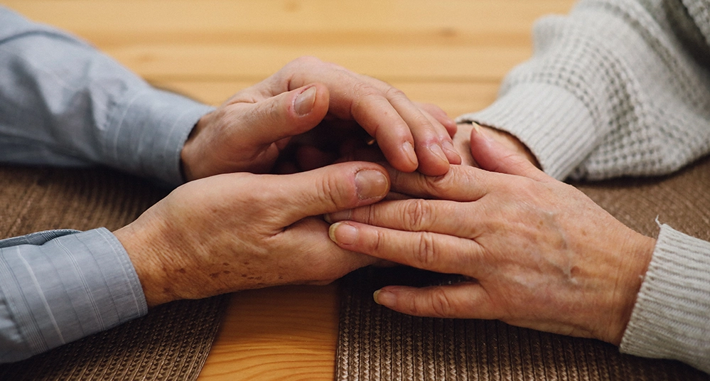 Two older people sitting across a table, hands lightly clasped
