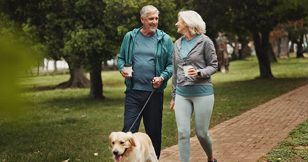 Managing Chronic Conditions After 65: Systems That Hold 3 Older couple walking their dog together on a park path