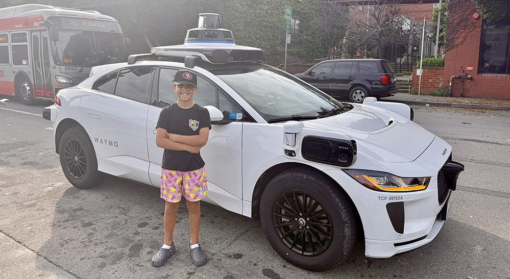 Boy in a San Francisco 49ers cap standing with arms crossed in front of a white driverless Waymo car