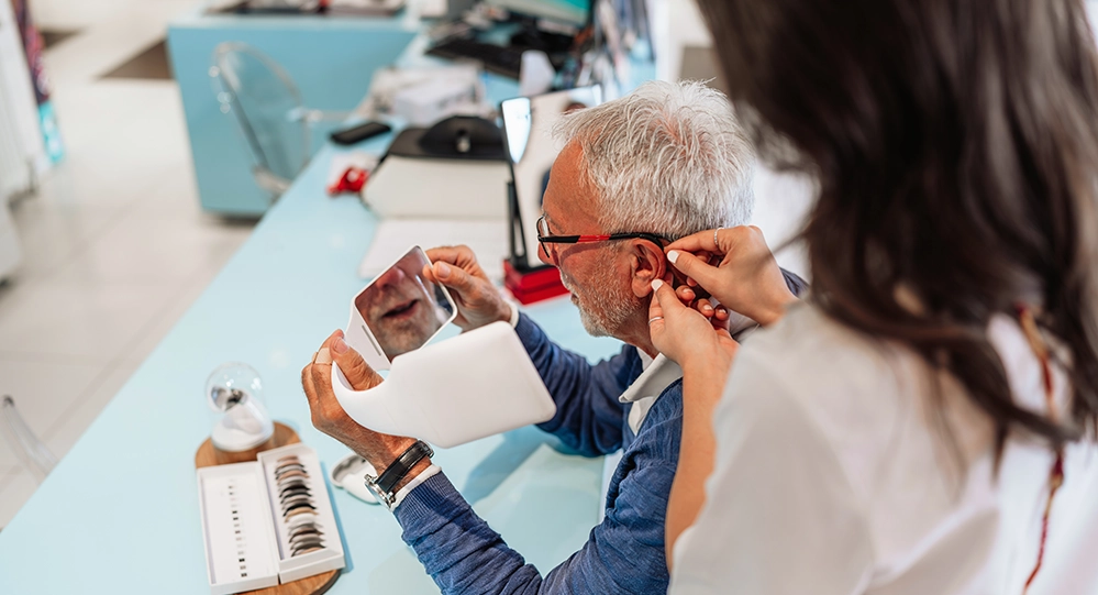 An audiologist fitting a hearing aid for an older man while he watches in a hand mirror
